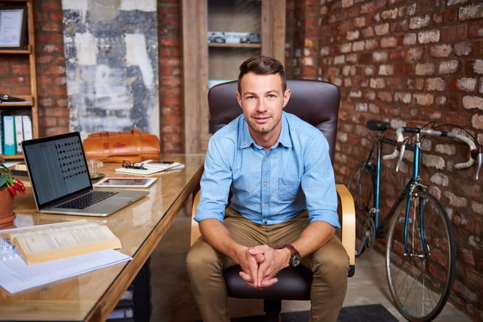A man seated in his home office with exposed brick walls, wearing a light blue shirt and khaki trousers. A wooden desk in front holds an open book, a laptop, a smartphone, and a leather bag. A bicycle is leaning against the wall in the background