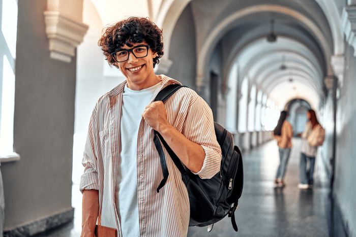 A young male student stands in a corridor. He is holding a rucksack and smiling at the camera.