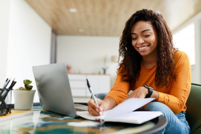 Young woman writing in a notebook with a laptop open beside her