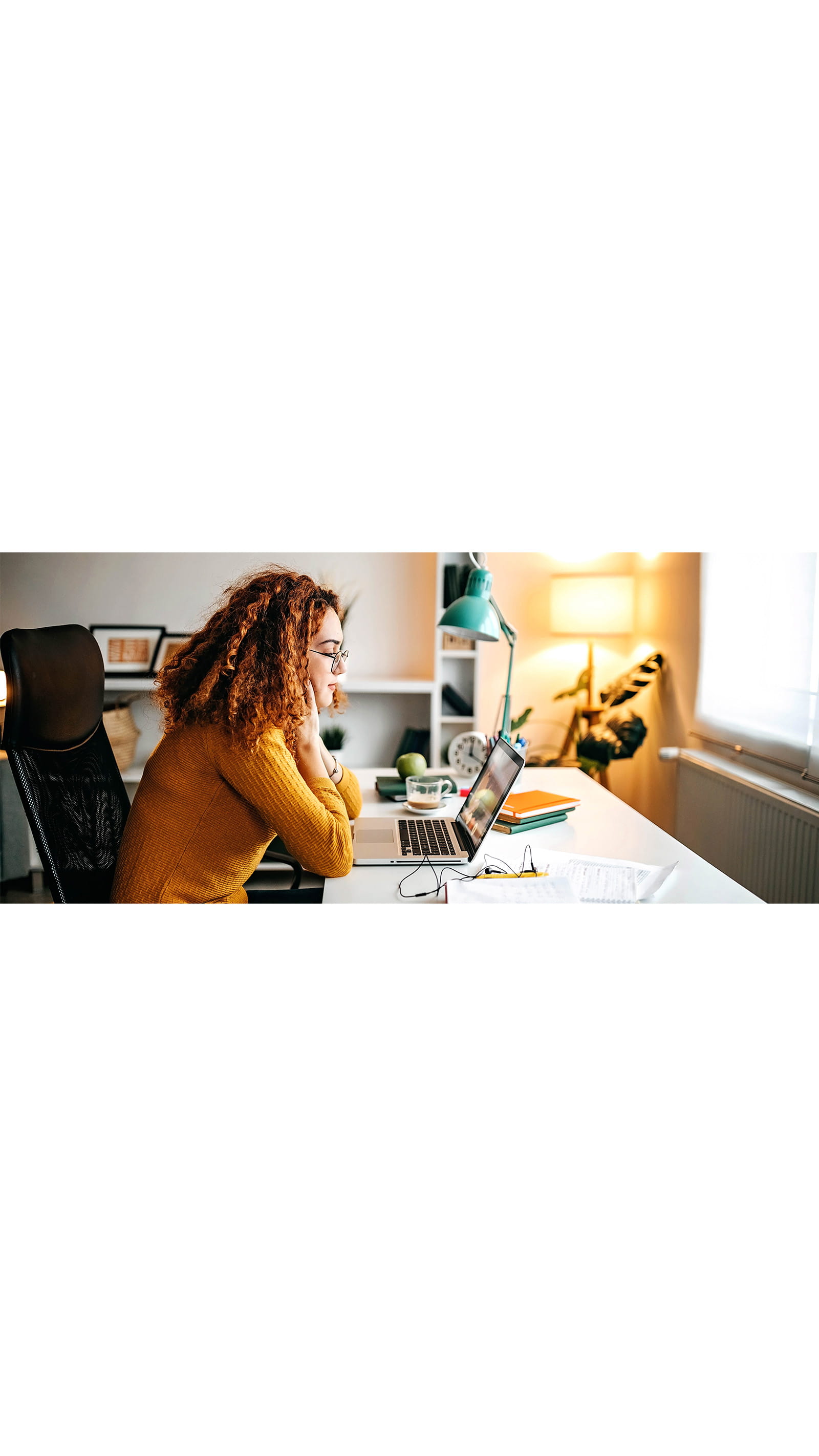 young woman student orange jumper curly hair laptop studying desk lamp