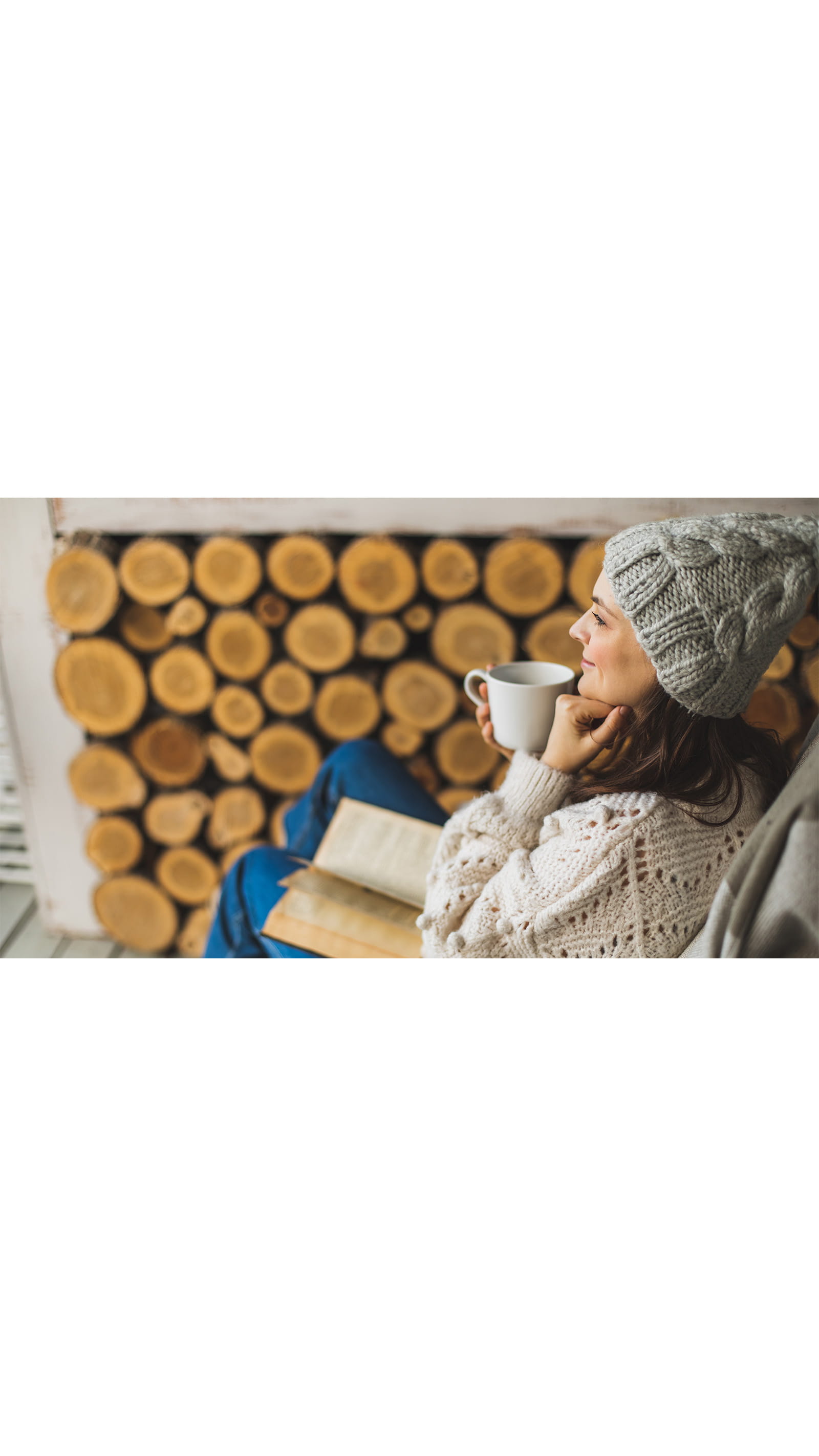 young woman knitted grey beanie hat cream jumper sweater mug hot drink book wood logs winter holiday