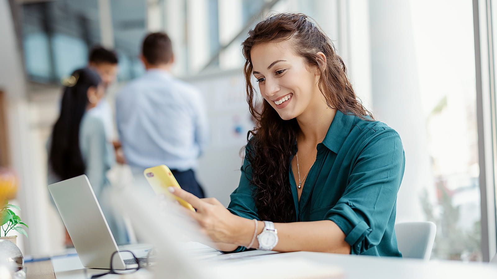 young woman employee working in an office smiling looking at mobile phone laptop desk