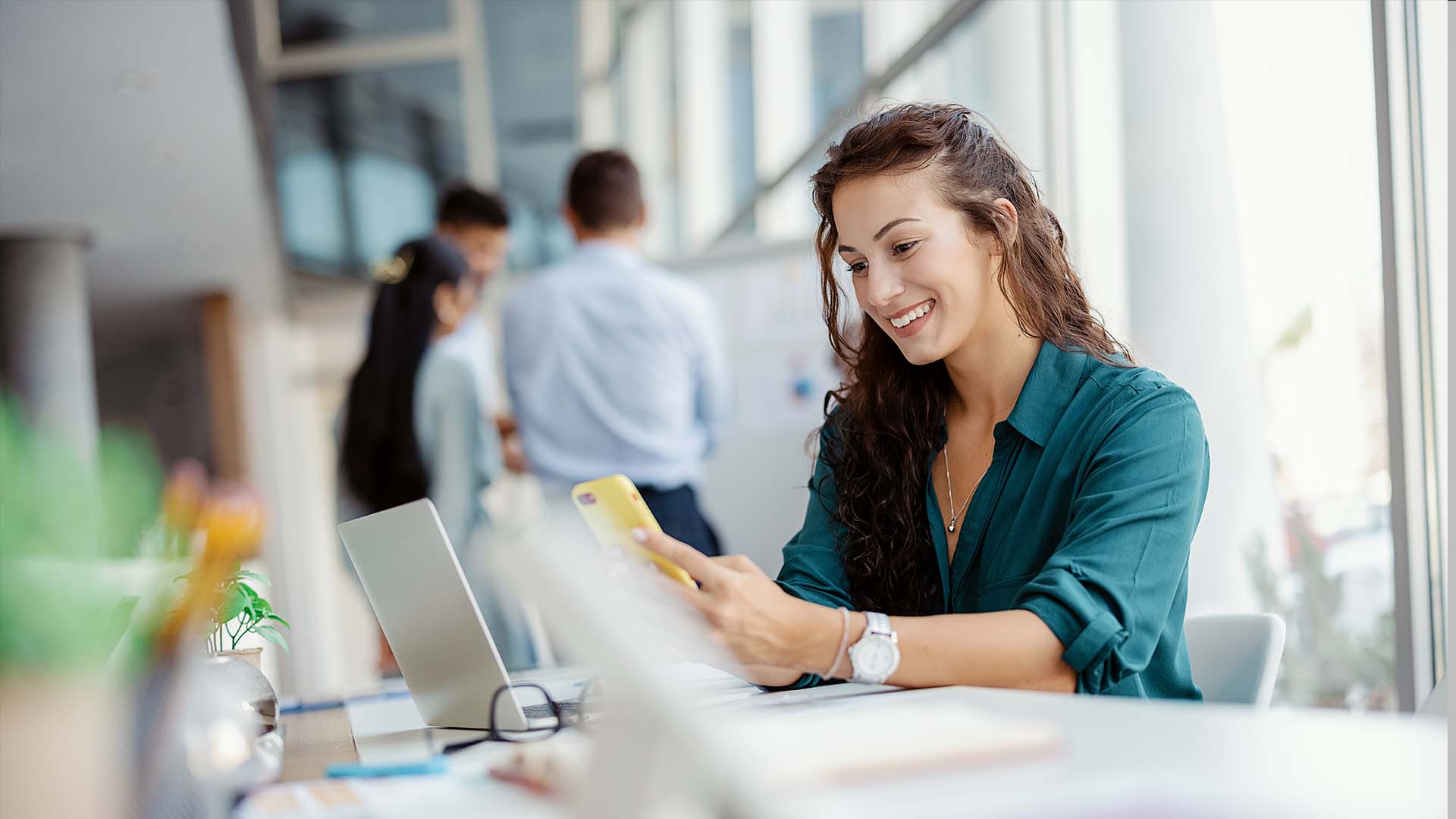 young woman employee working in an office smiling looking at mobile phone laptop desk