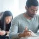 young students studying sitting in a classroom exams reading textbook turning page