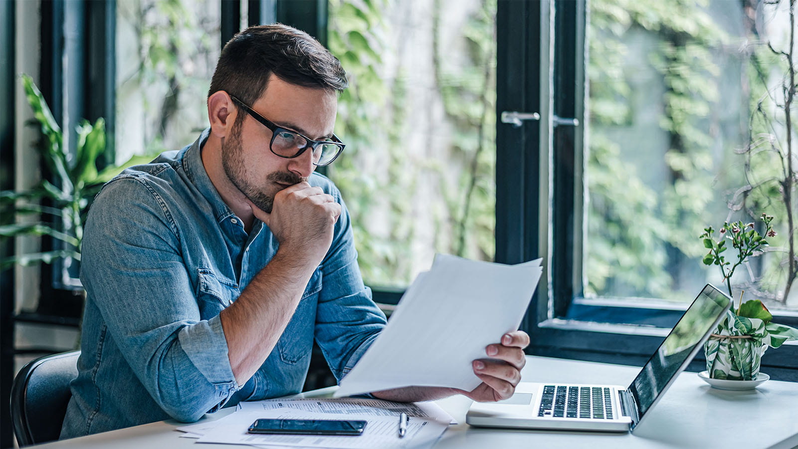 young man wearing glasses chin in hand looking at paperwork in an office desk laptop green plants