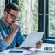 young man wearing glasses chin in hand looking at paperwork in an office desk laptop green plants