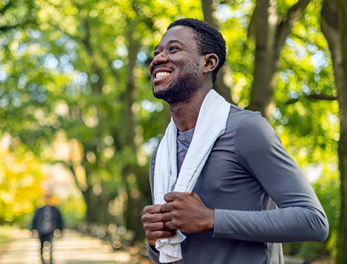 smiling young man jogger runner outside park green trees holding towel around neck