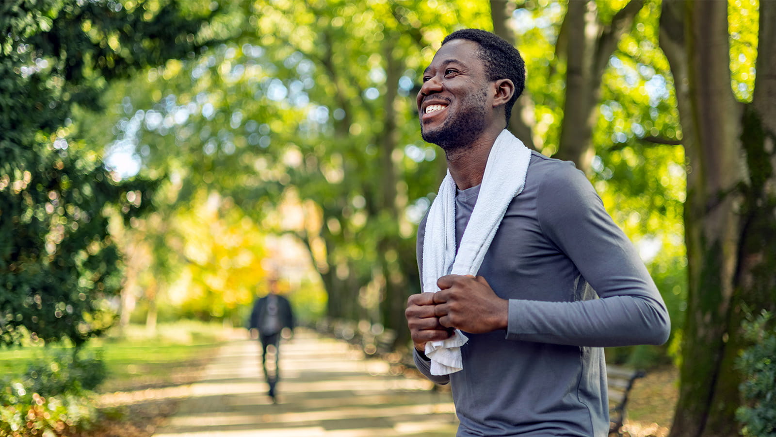 smiling young man jogger runner outside park green trees holding towel around neck