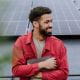 young man with a beard wearing a red shirt standing in from of solar panels on a rooftop sustainability