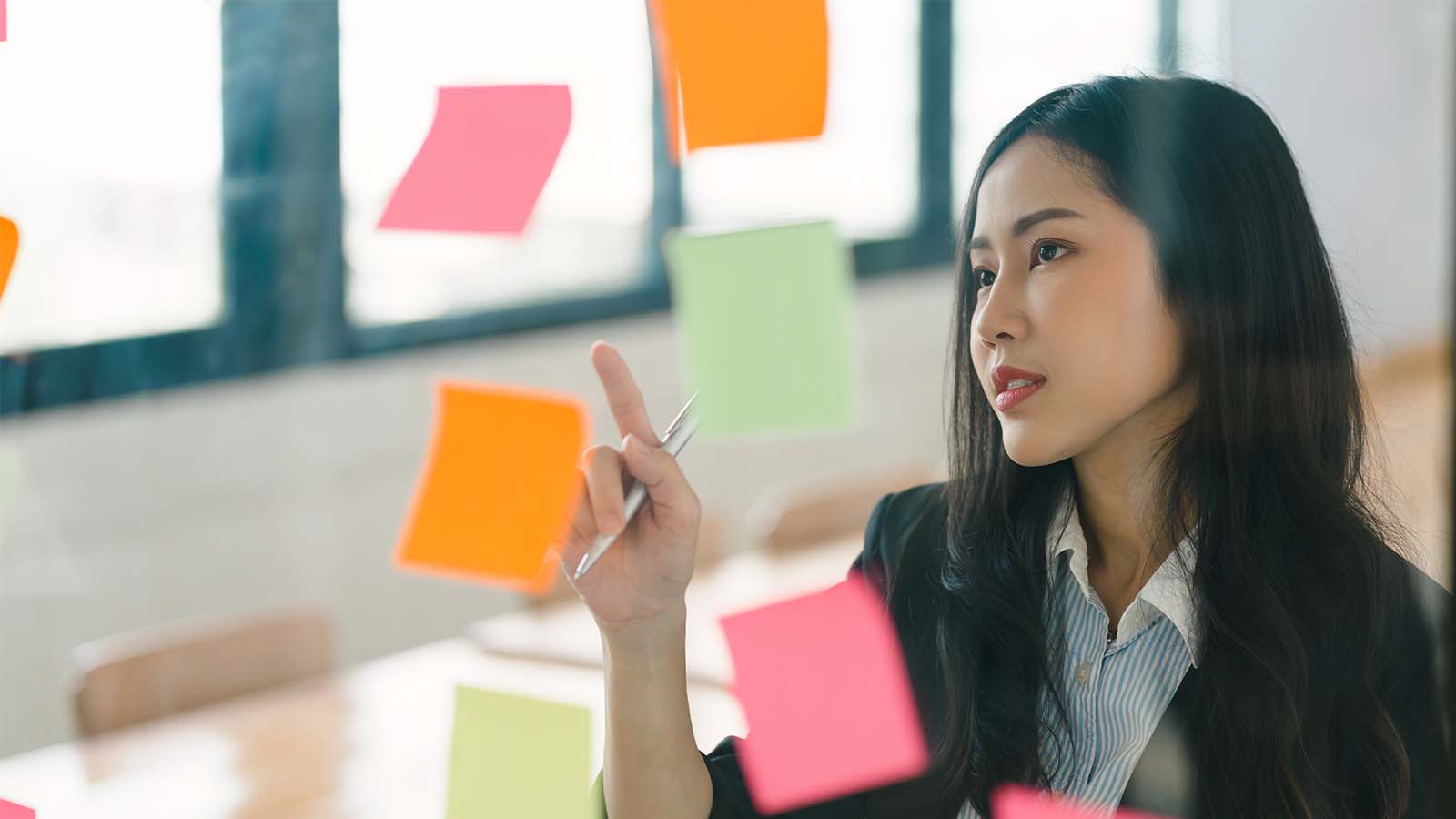 young woman employee working in an office meeting room pointing looking at post-it notes on glass