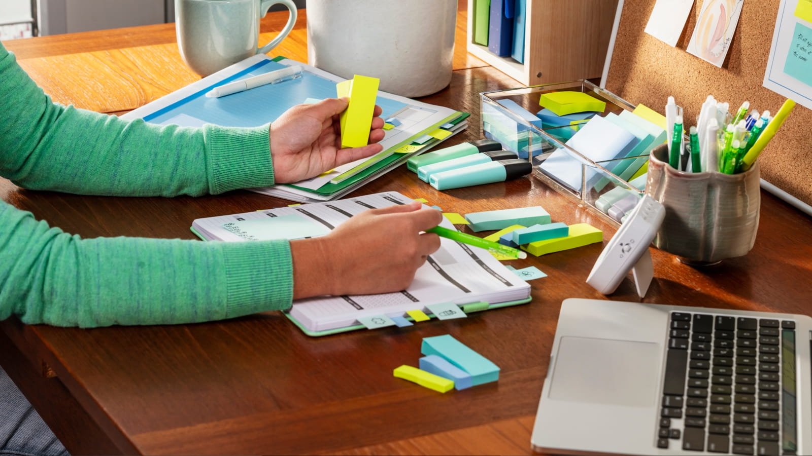 A person wearing a green sweater writes in an open planner at a wooden desk surrounded by colorful sticky notes, highlighters, pens, and a laptop.