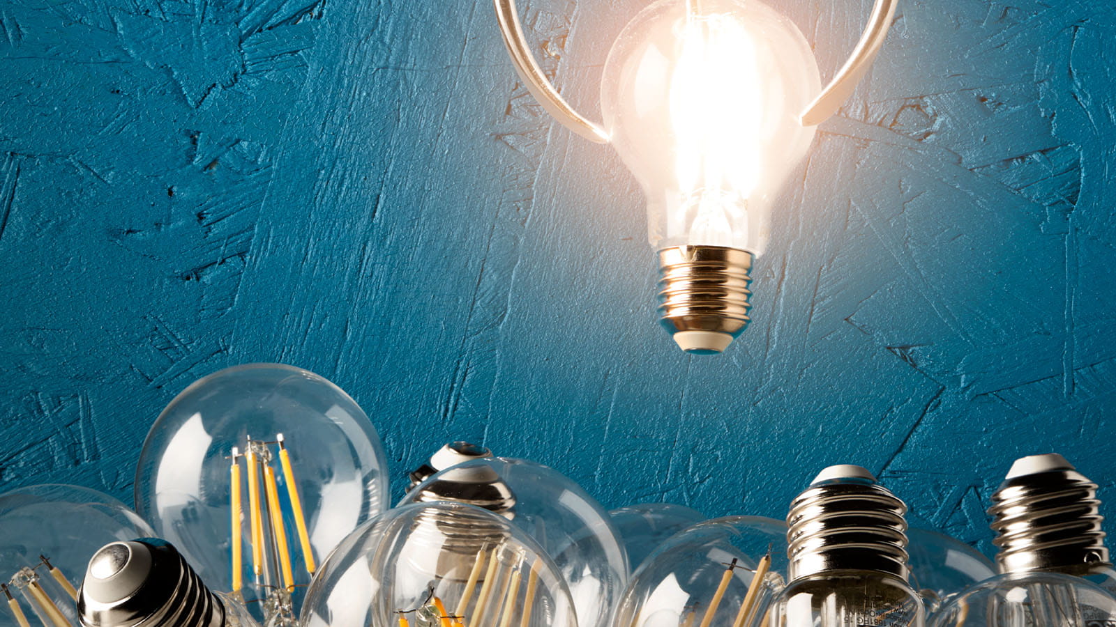 light bulb glowing being picked up by arcade claw above pile of many other filament light bulbs blue background