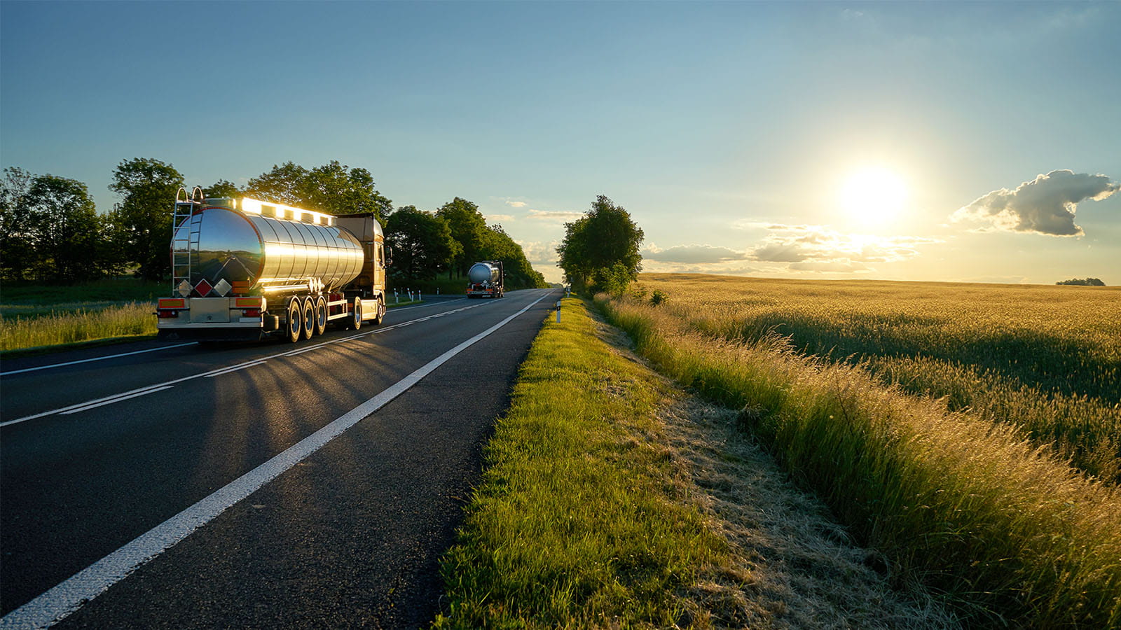 photo of an oil truck driving on a road at sunset sun setting over a field long grass blue sky