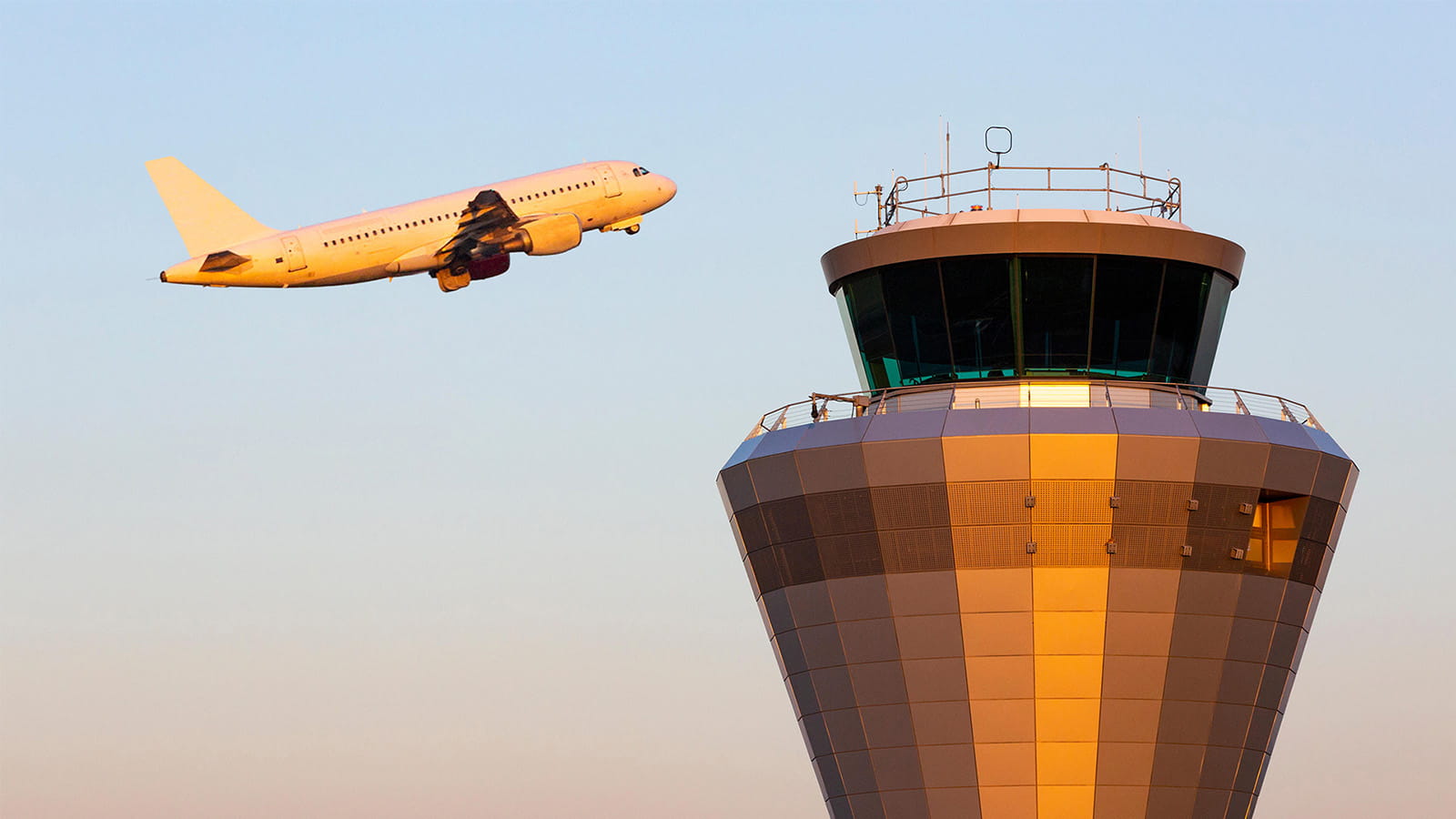 photo of an air traffic control tower building with a plane taking off behind orange sunset glow