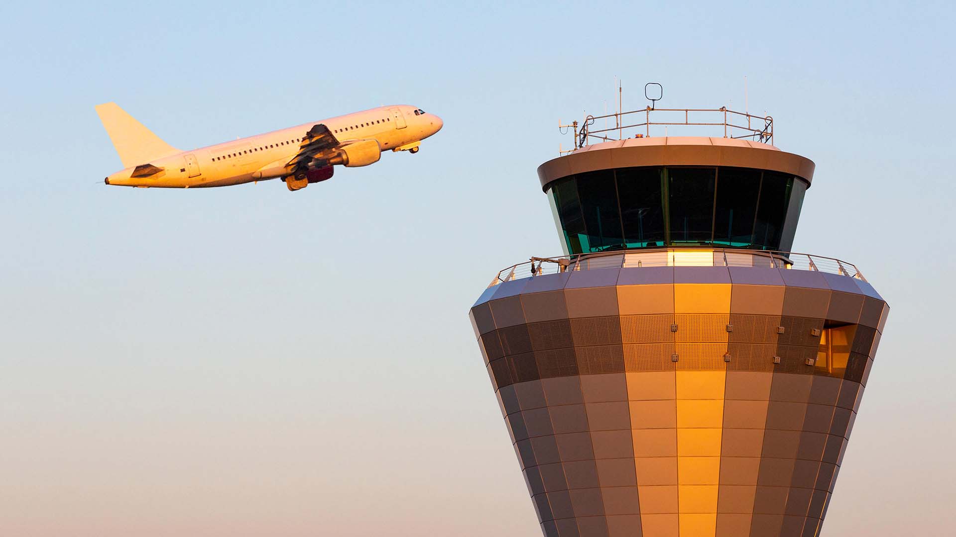 photo of an air traffic control tower building with a plane taking off behind orange sunset glow