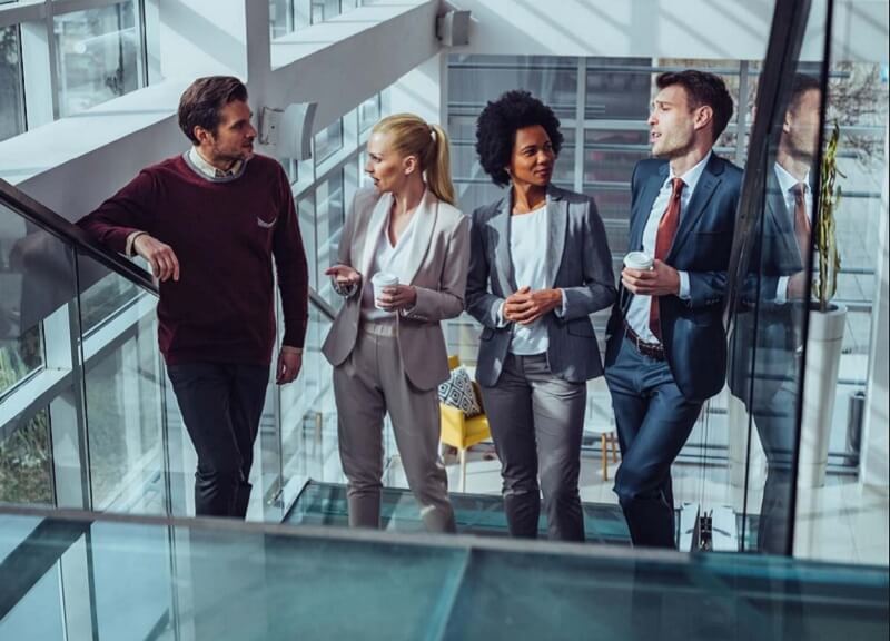 Four professionals walking together through a modern glass‑walled office corridor, holding takeaway coffee cups and talking as they move down a stairway.