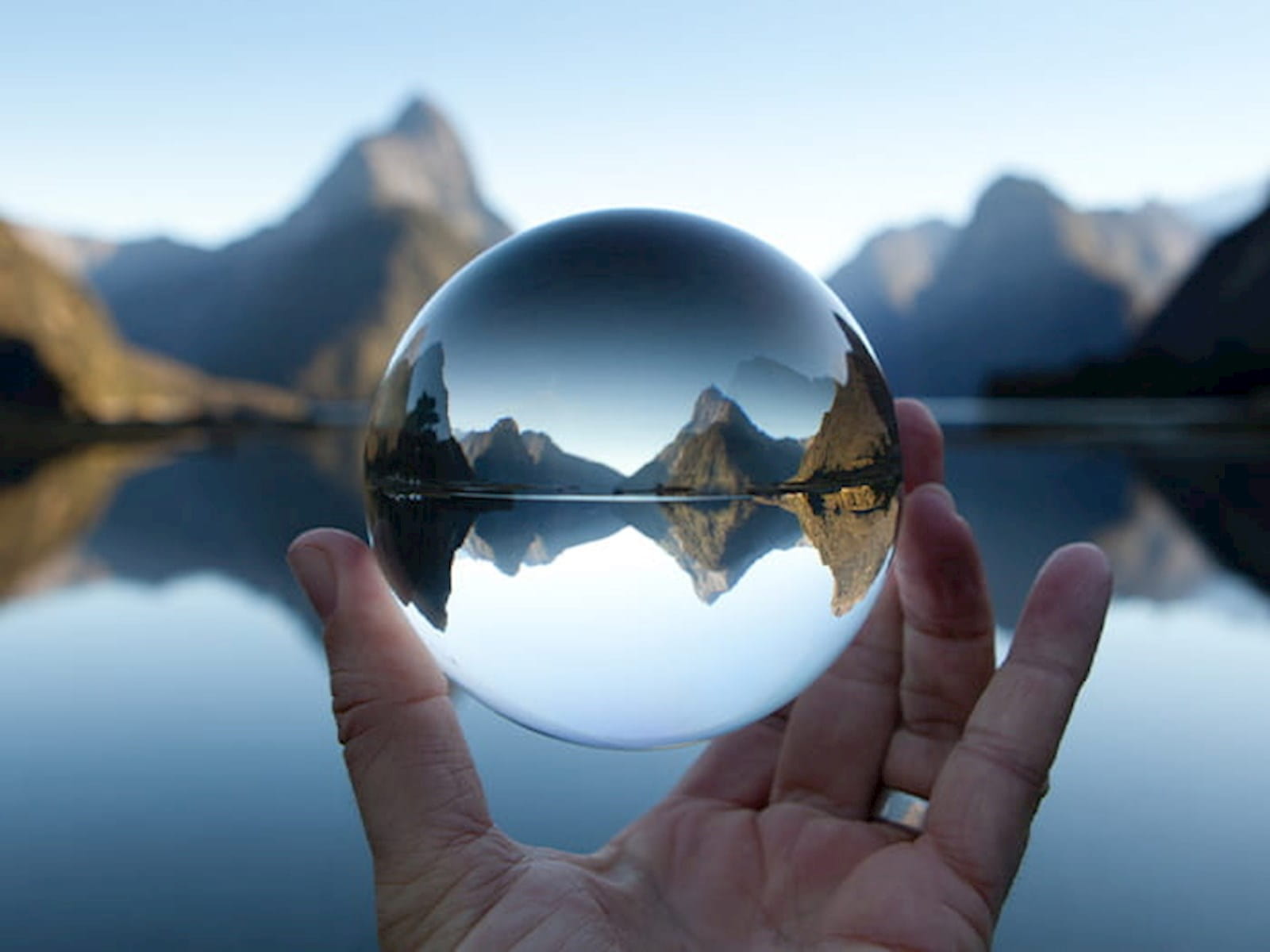 glass ball orb held in human person's hand fingers in front of lake and mountains blue sky water