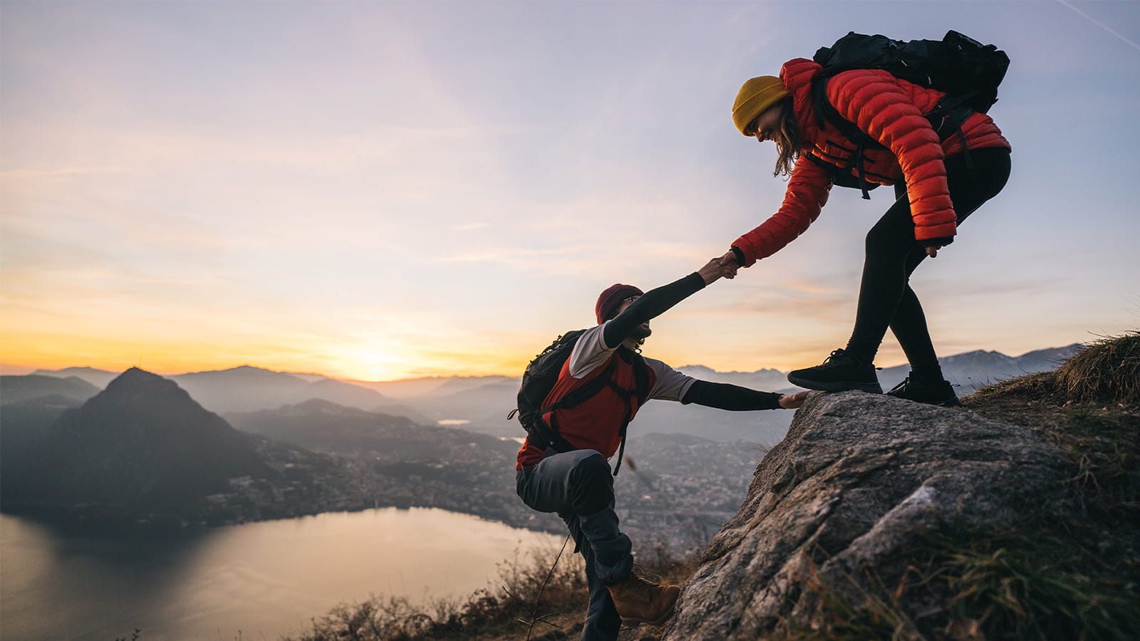 two hikers mountain climbers helping each other up to a peak rocks cliffs sunset