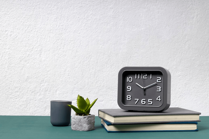 An analogue clock on top of two stacked, hardback book. Next to the books is a small potted succulent plant in a stone-like container and a grey cylindrical cup.