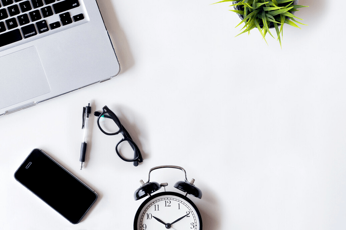 Overhead shot of a workspace featuring a laptop, a pair of glasses, a pen, a smartphone, an alarm clock and a small potted plant on a white surface.