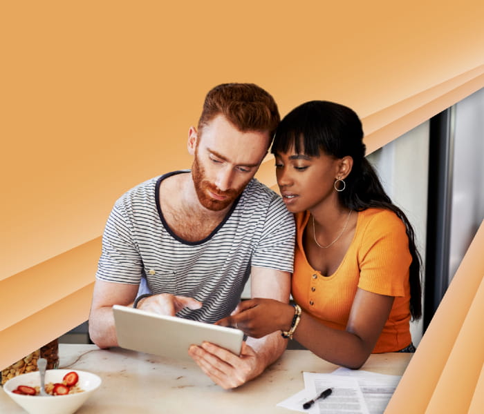 Man and woman sitting close to each other looking at a tablet the man is holding against an orange background