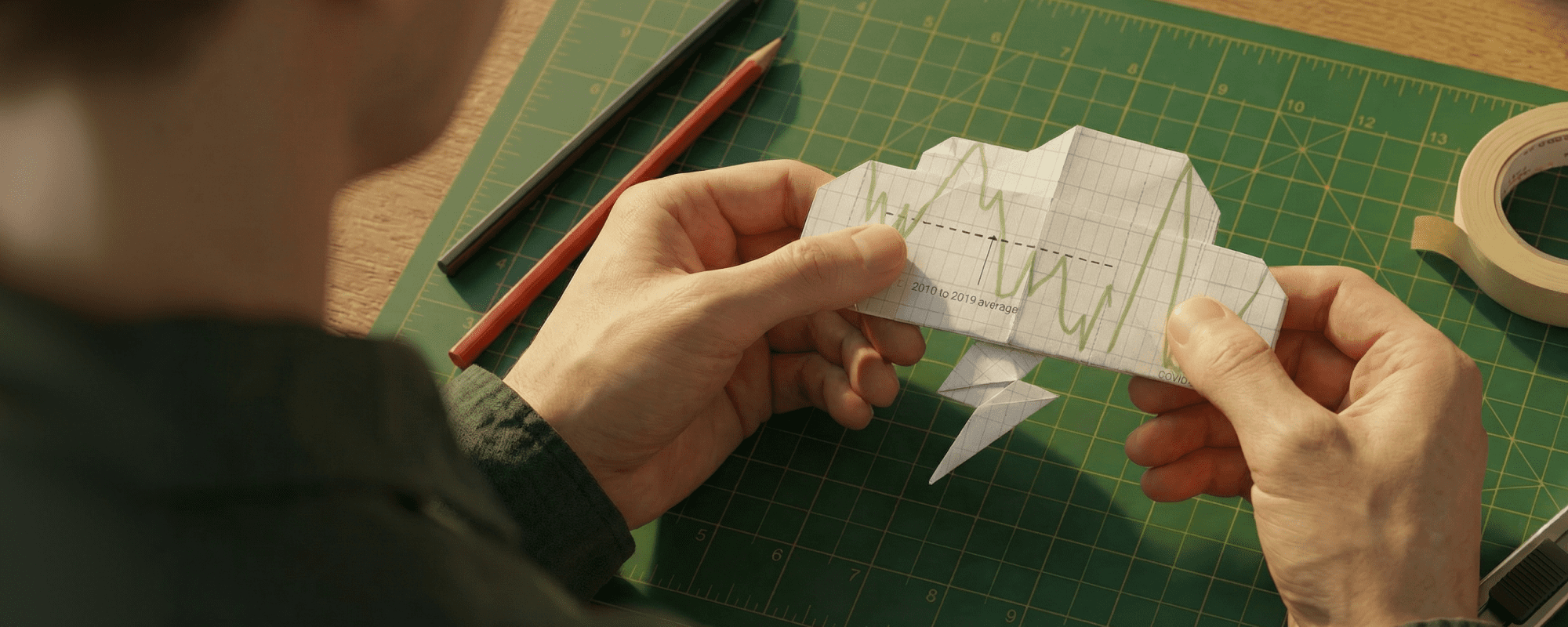 Hands holding a folded paper illustration with chart lines, photographed on a cutting mat.