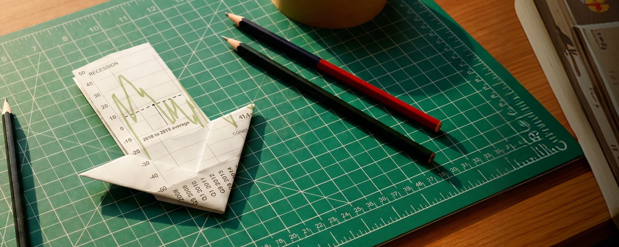 Origami downward arrow made from a financial chart on a green cutting mat beside pencils and a printed graph.