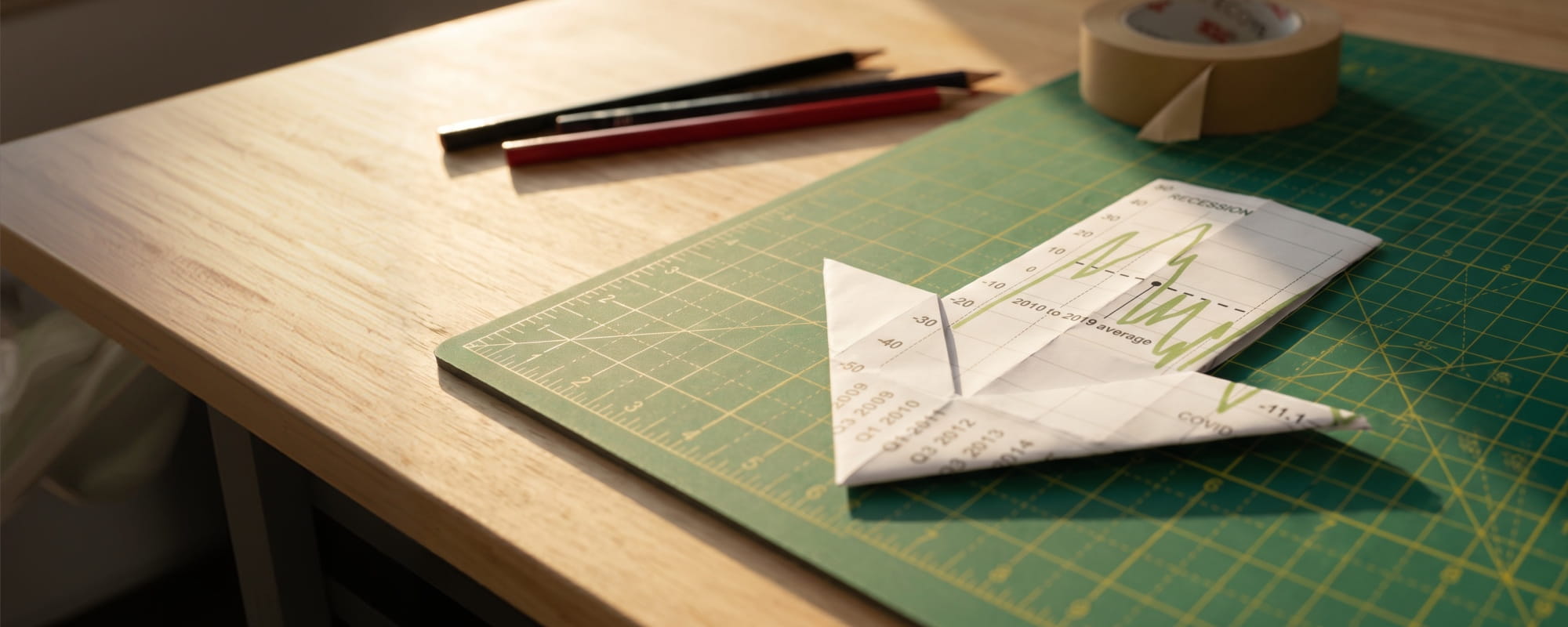 Origami downward arrow made from a financial chart on a green cutting mat beside pencils and a printed graph.