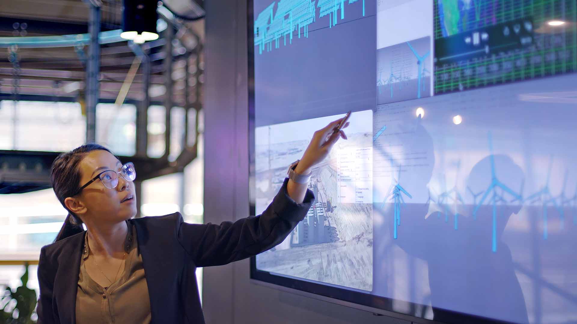 woman wearing glasses pointing at large screen showing data charts graphs office business