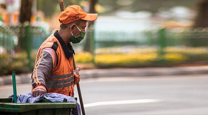 A bin worker holding a broom and pulling a wheelie bin along.