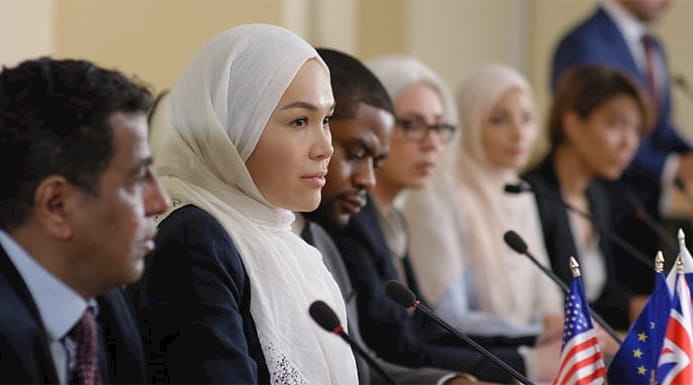 Woman in hijab sitting at table with multiethnic politicians answering questions during a press conference