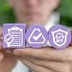 Three purple blocks held by a person, each showing a compliance-related icon: a clipboard labelled 'Compliance', a checkmark inside a triangle, and a shield with a checkmark