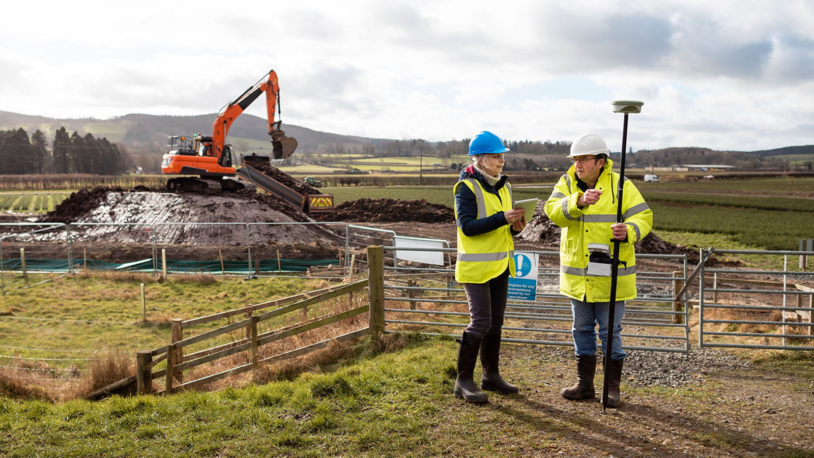 two construction workers surveyors wearing high vis hard hats in front of a work site with a digger