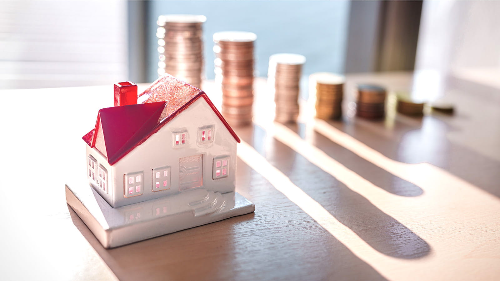 small model of a white house with red roof in front of piles stacks of coins money