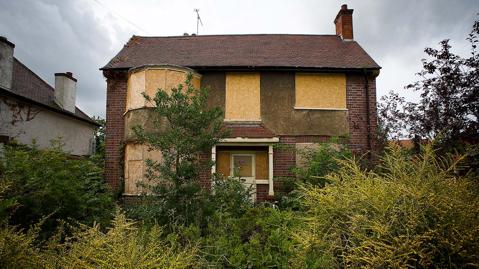 photo of an abandoned brick house two storey detached windows bordered up with overgrown garden foliage