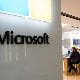 Interior view of a Microsoft store with a large glass wall displaying the Microsoft logo and colored square emblem, and people seated at wooden tables inside