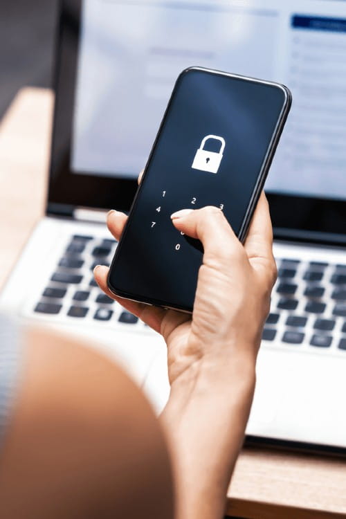 Close up of woman's hand holding a mobile phone, with a lap top open in the background. On the phone is the image of a padlock