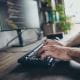 Close-up of hands typing on a keyboard at a desk with a large monitor displaying lines of code, suggesting programming or software development work.