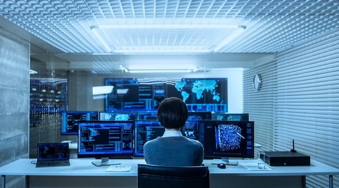A person seated at a desk in a modern control room, viewed from behind, facing multiple computer monitors displaying data, code, and world maps