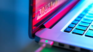 Close-up of a laptop keyboard and partially open screen with vivid pink and blue lighting reflections, and a connected cable visible near the hinge