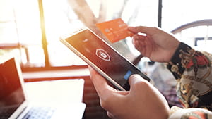 Close up of hands holding a smartphone displaying a security lock icon on the screen and a credit card in the other hand, with a laptop partially visible on a table in the background