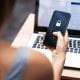 Woman holding a smartphone with a lock icon on the screen, indicating a security or password entry, in front of an open laptop displaying a webpage.