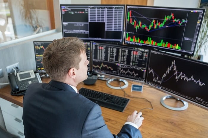 A man wearing a suit sitting at a desk with multiple computer monitors displaying stock market charts and financial data.