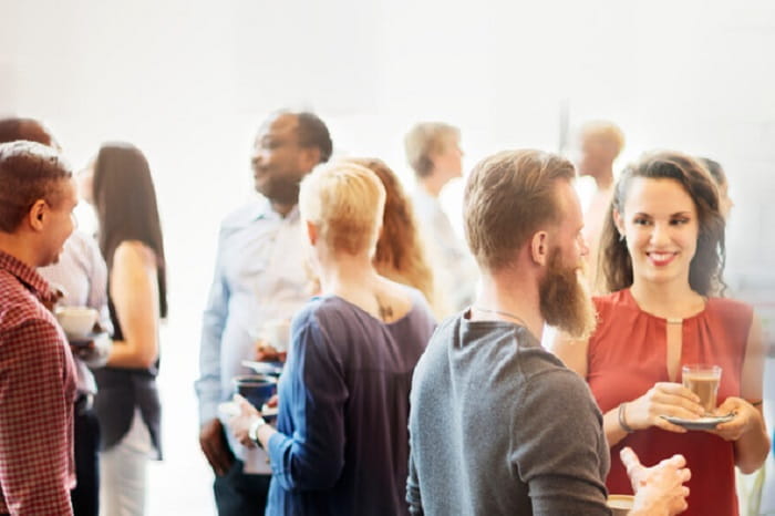 Group of people standing and conversing in a bright indoor setting, holding drinks and small plates, at a casual networking or social event
