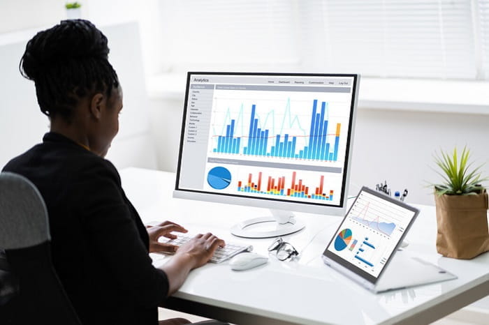 A woman working at a desk with a large monitor and a tablet displaying colorful bar charts, pie charts, and line graphs labeled “Analytics,” indicating data analysis or business reporting.