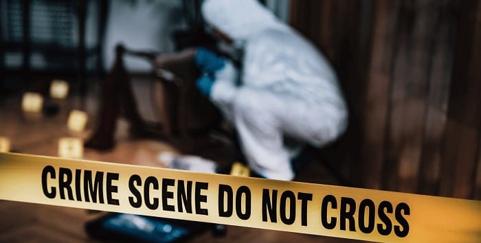 Tape with writing "Crime scene do not cross" cordoning off an area witha person wearing a forensic paper suit in the background.