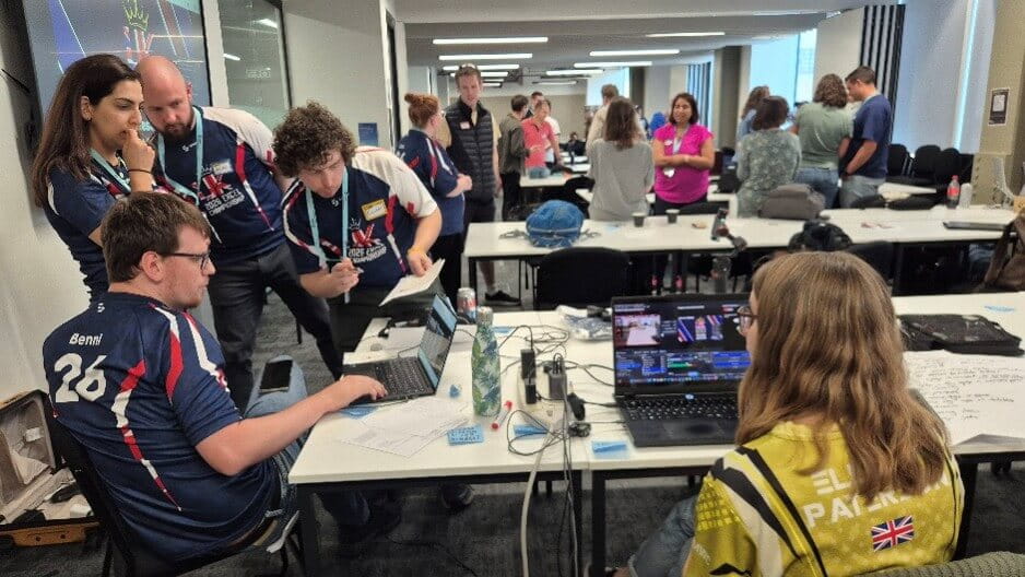 two men and a woman look at a seated man's laptop on a desk while another woman looks on