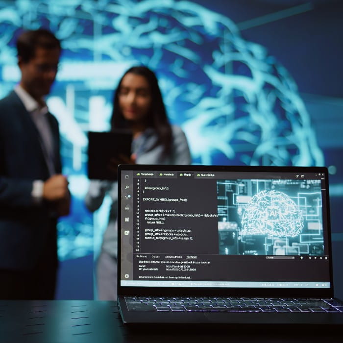 Laptop screen in foreground with digital visualisation of a brain. In the near background stand a man and woman in business clothes out of focus. 