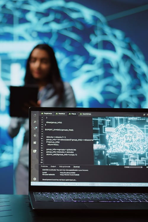 Laptop screen in foreground with digital visualisation of a brain. In the near background stands a woman in business clothes out of focus. 