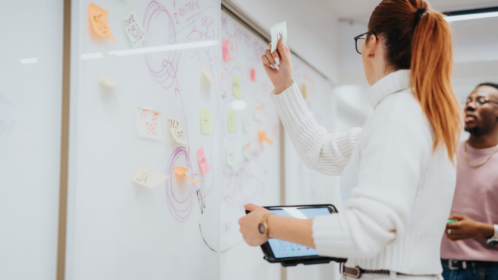 A woman adding a sticky note to the whiteboard while holding a tablet in her hand.